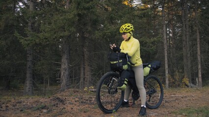 Naklejka premium The woman travel on mixed terrain cycle touring with bike bikepacking outdoor. The traveler journey with bicycle bags. Sportswear in green black colors. Magic forest park. Make a selfie smartphone.