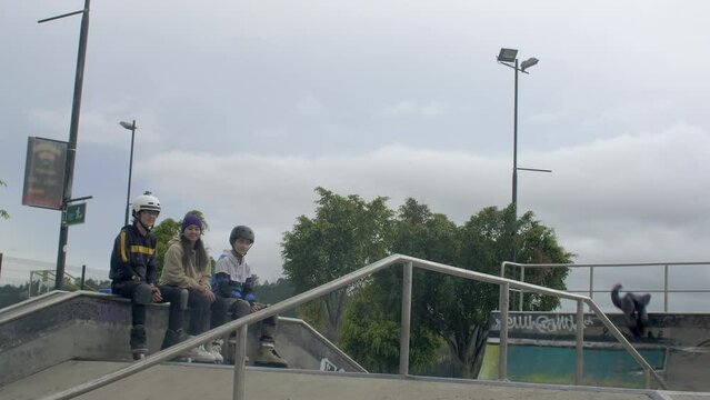 three males jump and spin at skatepark while three other people watch and clap at them 