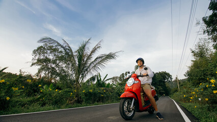 Man on red motorbike in white clothes drive on forest road trail trip. One men caucasian tourist go on scooter, nearby tropical palm tree. Asia Thailand ride tourism. Motorcycle rent Safety helmet.