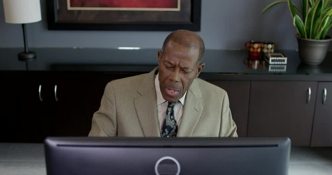 A serious older black businessman faces the viewer seated at a desktop computer and becomes increasingly more impatient while working. 35mm Medium Closeup Tripod Shot 4K.