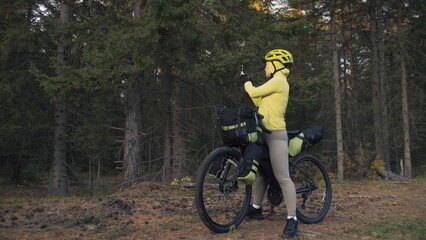 Naklejka premium The woman travel on mixed terrain cycle touring with bike bikepacking outdoor. The traveler journey with bicycle bags. Sportswear in green black colors. Magic forest park. Make a selfie smartphone.