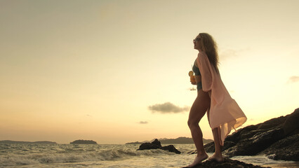 Silhouette woman stand on a reef rock stone in sea on golden sunset. Girl on tropical beach in green swimsuit and flutter in wind pink tunic silk shirt cape, drinks her orange cocktail Pina Colada