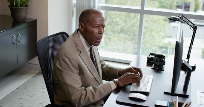 A serious black businessman sits at a desktop computer near a large office window and leans forward staring at screen with confusion as he works. 35mm Medium Closeup Tripod Shot 4K.