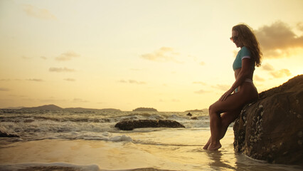 Silhouette sexy woman leaning on a rock reef enjoying and relax warm dark golden sunset. Woman in blue swimsuit and sunglasses. Concept rest tropical resort traveling tourism happy summer holidays