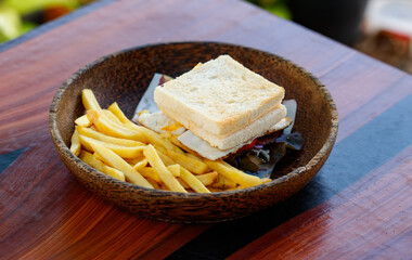 Closeup shot delicious tasty yummy homemade breakfast fried egg, pork bacon and cheese sandwich cutting edge bread with straight cut fresh frying french fries appetizer in wooden bowl on wood table