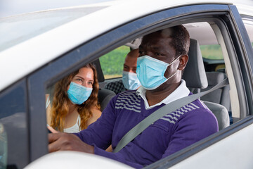 African american man in medical face mask driving car with friends in passenger seats. Concept of new life reality and health protection in coronavirus pandemic