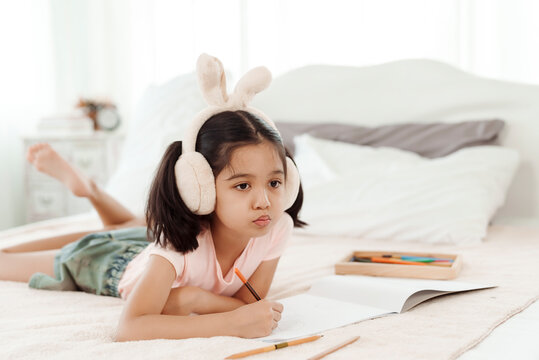 4k 50fps Cute little girl having fun in cute kindergarten age. Use colored pencils to draw a picture on the paper. Happily put the rabbit headphones on the bed. Concept family and happy learning