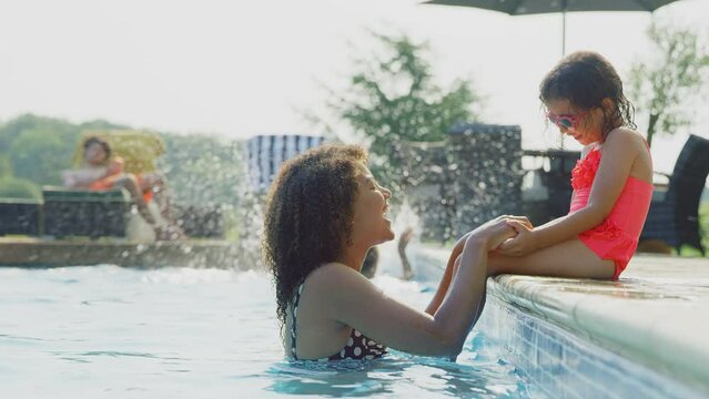 Mother having fun in outdoor swimming pool on summer vacation with daughter sitting on the edge wearing heart-shaped sunglasses - shot in slow motion