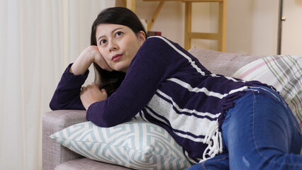 lazy Taiwanese girl is propping up on the couch and looking away into space with an unhappy...