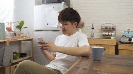portrait japanese young guy is watching online videos on his smartphone while sitting at dining...