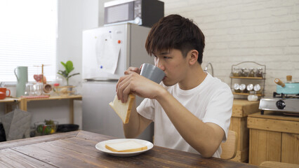 carefree young Asian man is biting into the toast and drinking coffee from cup while enjoying the first meal of the day on a tranquil morning at home.