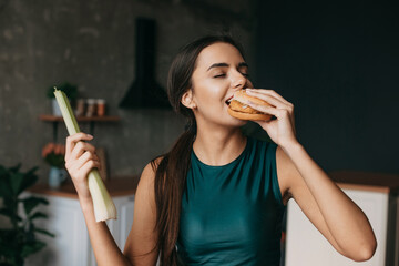 Fototapeta premium Caucasian sporty woman losing strength of will and bitting a cheeseburger. Beautiful portrait. Diet, vegetarian food. Junk food.
