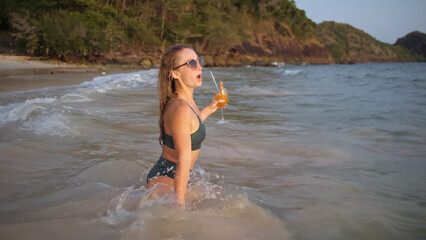 Attractive woman stand knee-deep in sea on golden sunset. Girl on tropical beach in green swimsuit having fun and waving his hands, drinks her orange cocktail Pina Colada. Big waves hitting legs