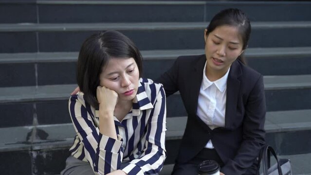 Asian Woman Wearing Business Suit Is Comforting Her Depressed Colleague By Patting Her On The Back As She Is Complaining On An Outdoor Staircase.