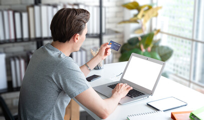 Young man using credit card, shopping online or ordering via Internet. Laptop with blank white screen and tablet. Man working at home office, ordering products online.