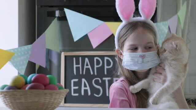 Portrait of funny Easter kid in bunny ears with cat. Pretty little girl having fun in springtime in decorated room. Cute child wearing protective face mask