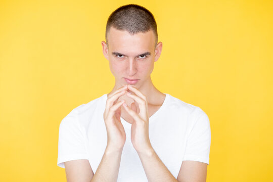 Sarcastic man. Evil look. Bad intention. Conspirator mockery guy in white t-shirt holding hands together showing sardonic smile isolated yellow.