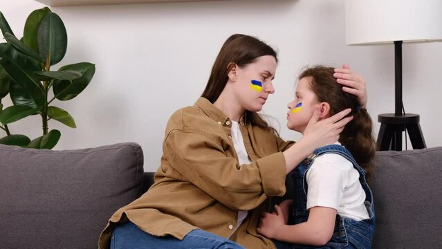 Loving Ukrainian young mother hugging small upset daughter with flag of Ukraine on face, family sitting on sofa at home, poverty concept. Crisis, peace, stop aggression, child against Russian war