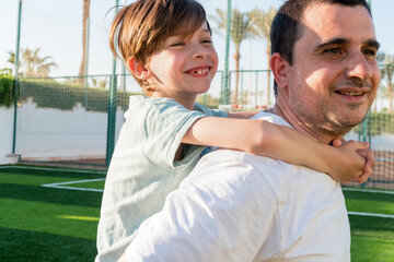 Side view of smiling father carrying cheerful boy on back while spending time together at football...