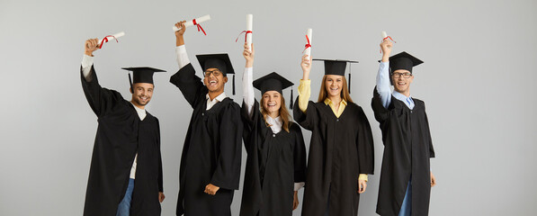 Portrait of joyful successful multiracial graduates with scrolls of diplomas in hands on gray...