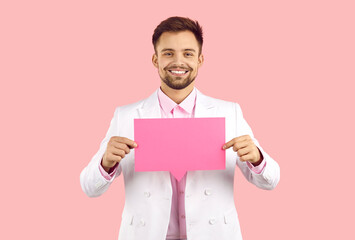 Attractive smiling man holding blank pink speech bubble while standing on pastel pink background. Young caucasian man in white formal suit holding paper layout with empty space for text. Copy space.