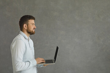 Portrait in profile of a young and confident man standing on a gray background with a laptop in his hands. Smart caucasian male programmer or office worker looking towards a free space for text.