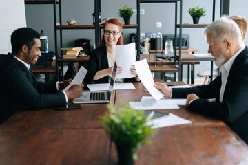 Cheerful female team leader in glasses holding corporate meeting with multiethnic colleagues in boardroom sitting at desk. Positive businesswoman discussing project with business partners in office.
