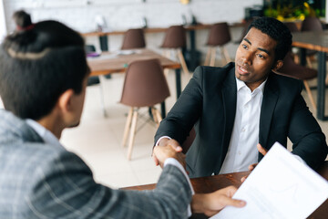 Close-up to two satisfied multiracial businessmen shaking hands over desk after successful negotiations, closing sealing deal, sitting at desk in modern office room. Concept of interracial cooperation