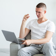 Small gesture. Begging man. Online conference. Gloomy casual guy sitting sofa looking laptop showing little size with fingers in light room interior.