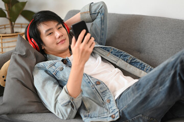 Obraz premium Handsome asian man wearing headphone resting on couch and using smart phone.