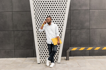 Handsome successful businessman with yellow laptop standing near the office, posing for the photography. Pensive african american male freelancer looking at side, lost in thoughts about new project