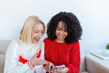 Happy smiling young women friends looking at photos on mobile phone. Girls havin fun. Two Female Friends Relaxing At Home Talking Together