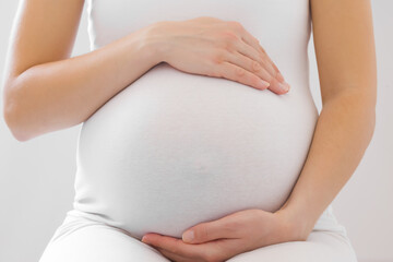 Young adult pregnant woman in white clothes touching big belly and feeling her baby move on light gray background. Pregnancy time concept. Expectation time. Closeup. Front view. - obrazy, fototapety, plakaty