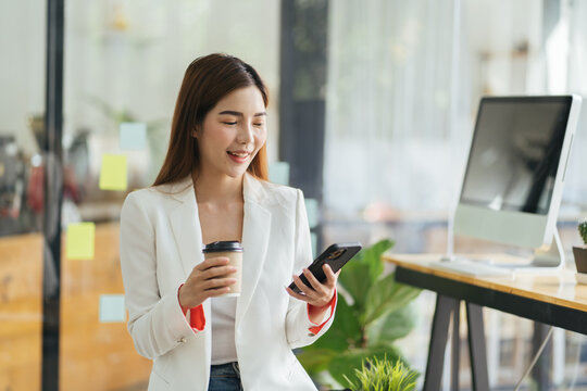 Asian business woman in cafe using mobile phone. Girl chatting on smartphone and drinking coffee in restaurant near window, smiling at cellphone screen.