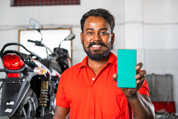 Fototapeta na wymiar Mechanic showing green screen mobile phone by looking at camera at workshop - concept of advertisement, promotion, online booking for maintenance and repair service.