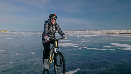 Woman is riding bicycle on the ice. Girl is dressed in a silvery down jacket, cycling backpack and helmet. Ice of the frozen Lake Baikal. Tires on bike are covered with spikes. Traveler is ride cycle.