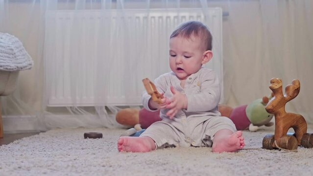 A happy child is playing with a rattle on the carpet in the nursery. Toddler baby boy is playing on the floor with a wooden toy, age eight months