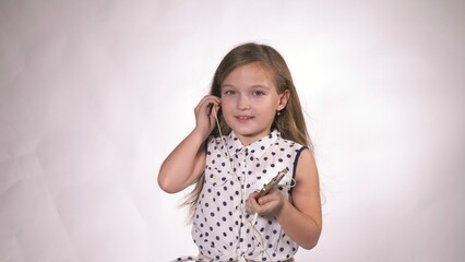 Fototapeta premium Child with earflaps dancing at studio background. The girl listens to music on the smartphone. The kid has loose long hair. Beautiful cute happy little girl listens to music on Headphones.