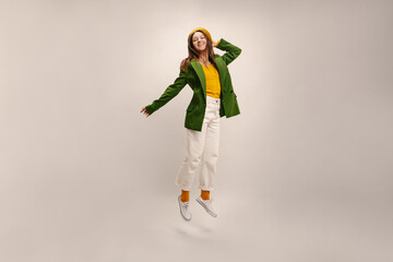 Happy young caucasian woman in beret, jacket and trousers is jumping in studio on white background. Brunette has fun at photoshoot. People sincere emotions lifestyle concept.