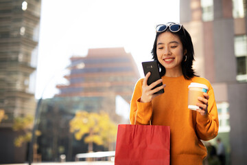 Pretty chinese girl using mobile phone and holding coffee to go. Beautiful woman enjog in coffee while walking on the city street.