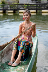 Young girl paddling on a wooden canoe in traditional clothes, and Hindu temple surrounded by body of water.