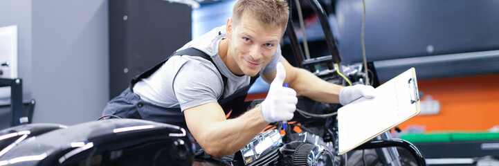 Young car mechanic is diagnosing a motorcycle and holding clipboard and thumbs up