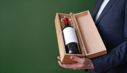 Male hands holding wooden gift box with bottle of red wine.Unrecognizable man offering,advertising alcohol drink on green background. Copy space