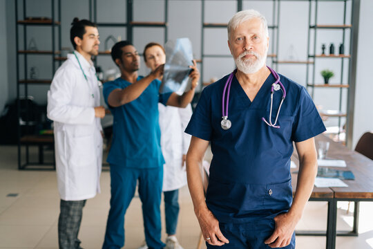 Portrait of serious mature older surgeon in blue uniform standing in medical meeting office, looking at camera. Multi-ethnic team of physician working in background, discussing MRI scan of patient.