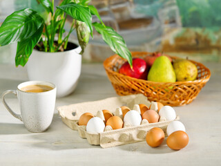 Brown and white chicken eggs in an open cardboard box. preparing breakfast in the morning sun in a kitchen table. mug of coffee, fruits and flowers on the table near the window. easter eggs Close up