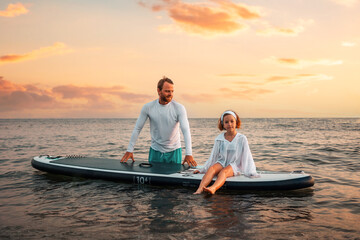 Father and daughter surfing on a sup board. Sea and sunset sky at the background. Summer vacations....
