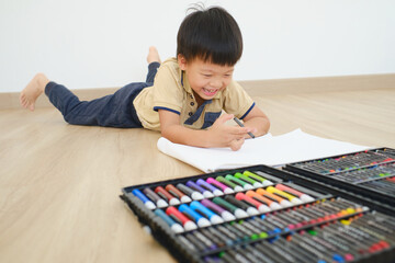 Cute little Asian kindergarten boy child lying on his stomach on the floor while drawing with colorful crayons and lot of painting tools at home, Creative play for kid, improve focus in child concept