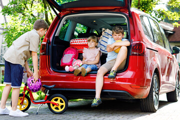 Three children, two boys and preschool girl sitting in car trunk before leaving for summer vacation with parents. Happy kids, siblings, brothers and sister with suitcases and toys going on journey