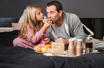 daughter gives muffin to father eating breakfast in bed
