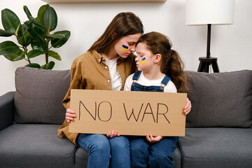 Ukrainian caring young mother and unhappy small daughter kid sitting on grey sofa holding banner...